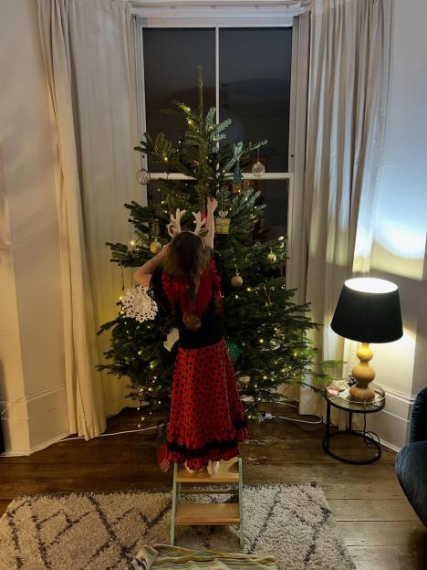 A child in a red and black dress and reindeer antlers stands on a stool decorating a Christmas tree.