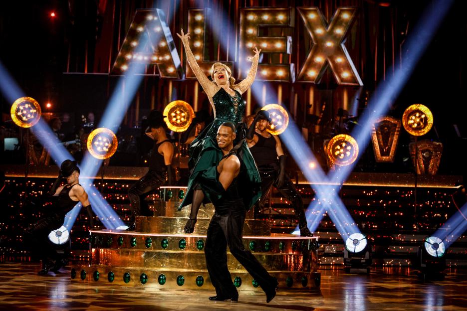 Alex Kingston in a green dress being carried by Johannes Radebe on a stage with "ALEX" in the background.