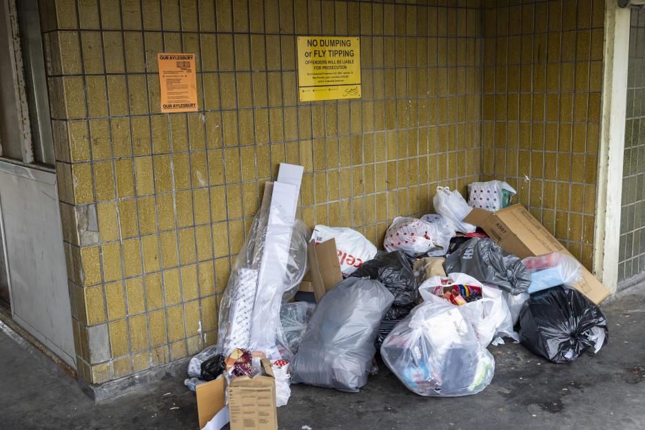 A pile of household waste on the ground next to a tiled wall, with a "No Dumping" sign above it.