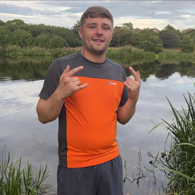 A man with short brown hair and a beard, wearing an orange and grey shirt and dark shorts, stands by a lake and forms "rock on" hand gestures.