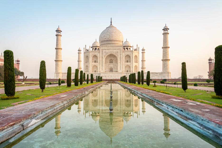 Taj Mahal monument at sunrise in Agra, India, reflected in a long pool of water.
