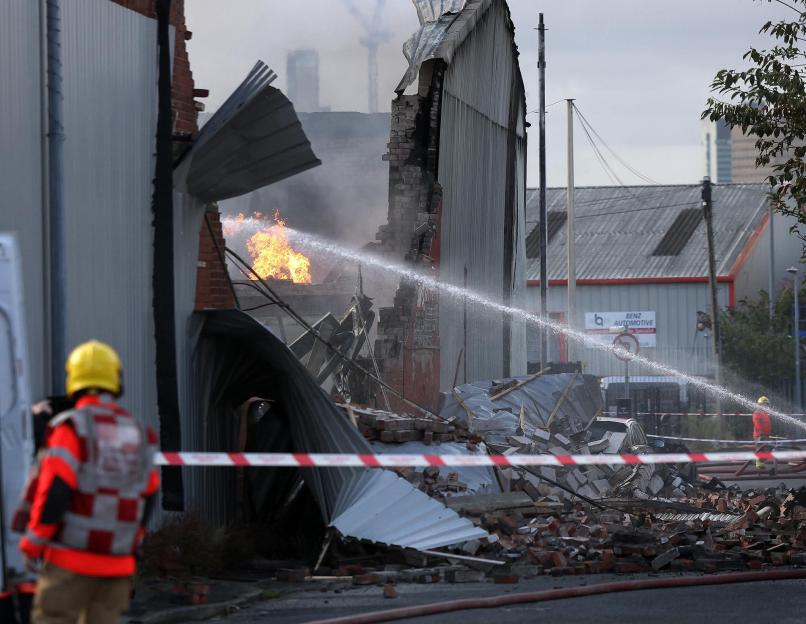 Firefighters spray water on a burning commercial building, its brick and corrugated metal walls collapsed, with debris scattered on the ground.