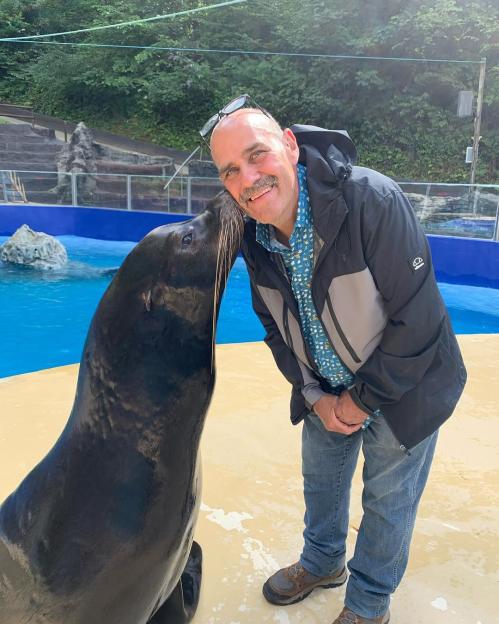 A man smiling as a sea lion nuzzles his face.
