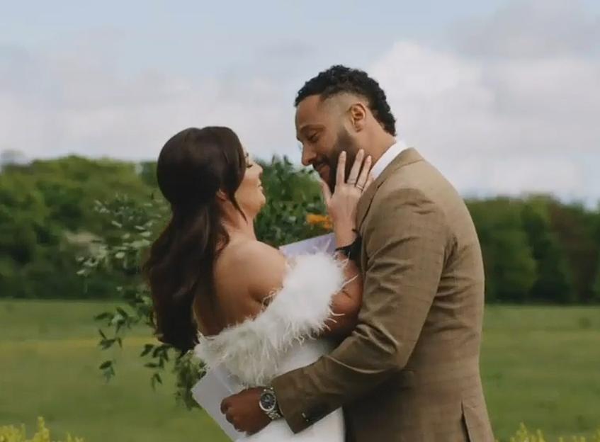 A couple embracing outdoors, with the woman in a white feathery dress touching the man's face.