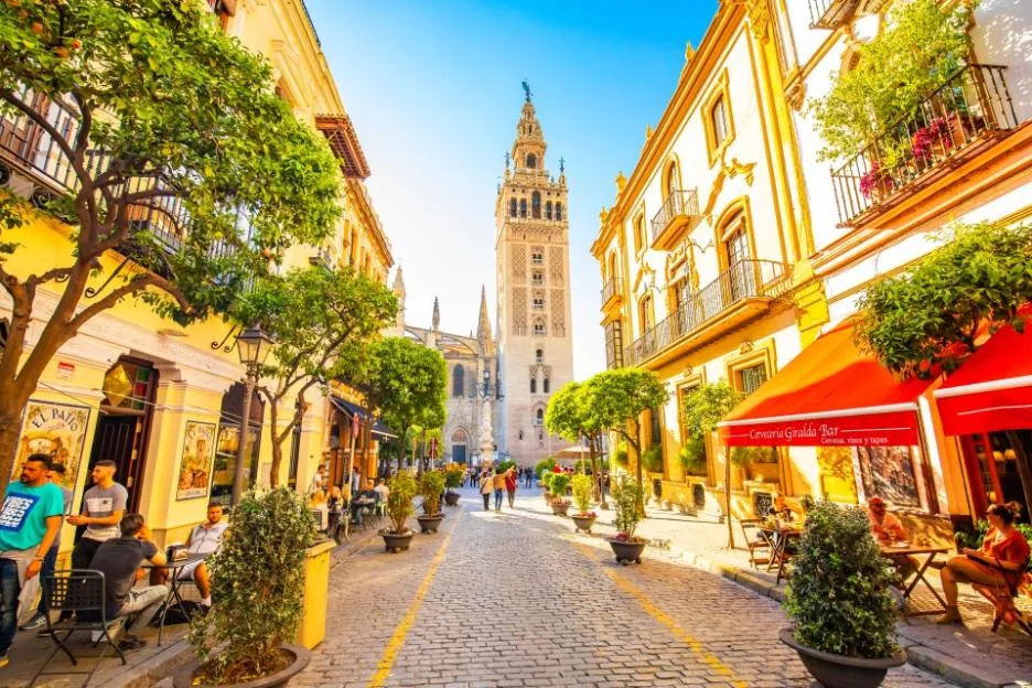 A sunny pedestrian street in Sevilla, Spain, flanked by yellow buildings, outdoor restaurants, and green trees, with the Giralda tower visible in the distance.