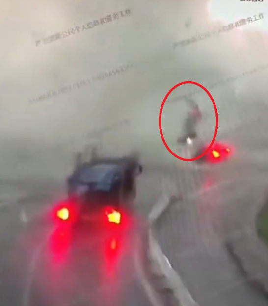 A blurry, high-angle image of a car with its tail lights on and a figure on a bicycle, circled in red, on a wet street.