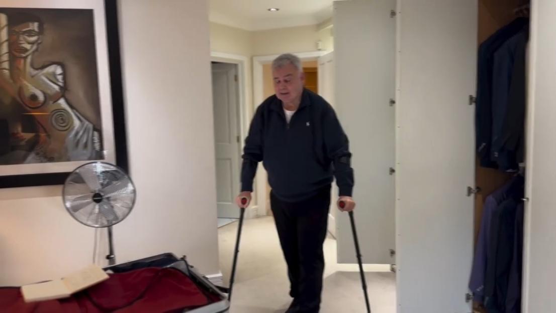 A man with crutches walks in a hallway, past a large framed picture of a woman's torso, a fan, and an open closet.