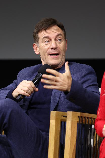 Jason Isaacs speaking at a panel, holding a microphone.