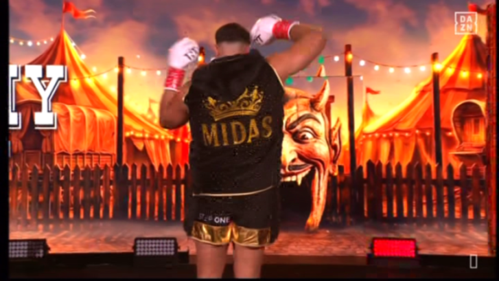 Boxer Tommy Fury wearing a robe with "MIDAS" and a crown on the back, standing in a carnival-themed setting.