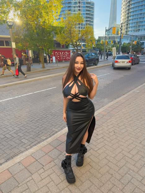 Louisa Khovanski in a black leather dress with cutouts and a high slit, standing on a city sidewalk.