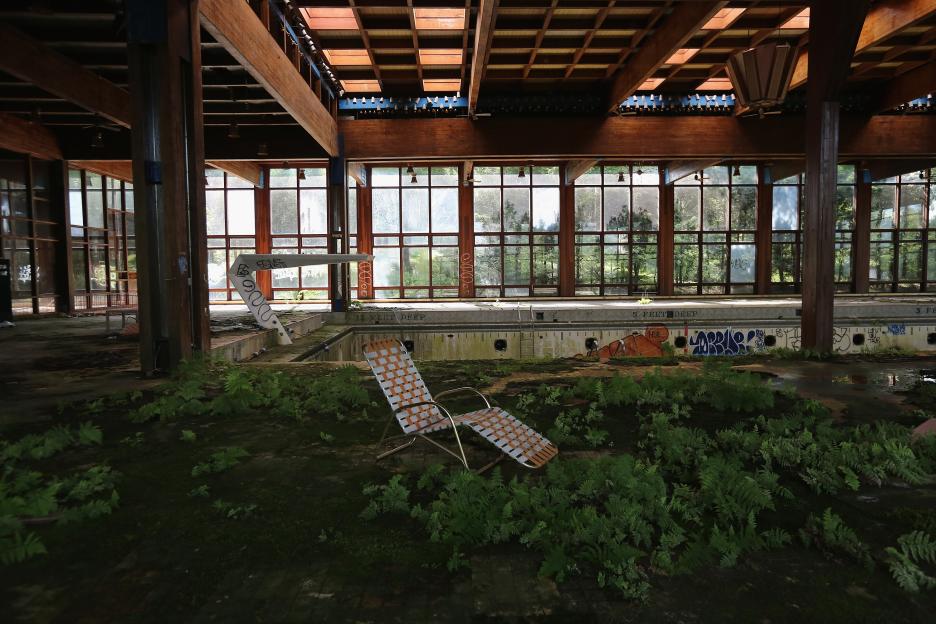 An abandoned indoor swimming pool with a diving board, a deck chair, and an empty pool, all covered in overgrown ferns and graffiti.