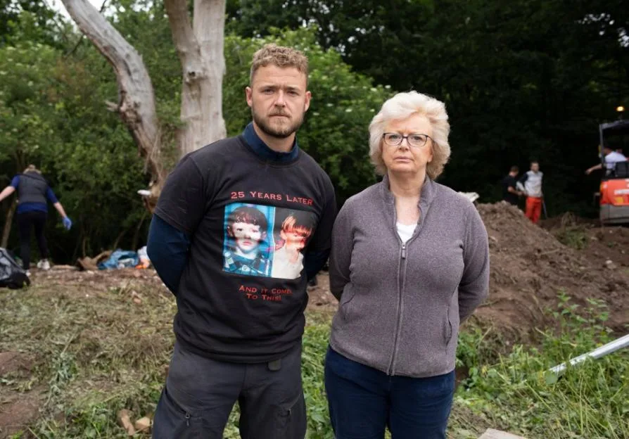 A man and a woman standing outside, the man wears a shirt with a photo of two children and the text "25 Years Later, And It Comes To This!"
