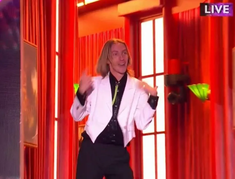 A man with blonde hair and a white suit dances in front of red curtains and a window.