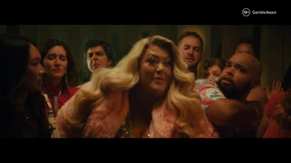 A crowd of diverse people, with a blonde woman in a pink fur coat and sequin dress in the foreground, all looking intently at something out of frame.