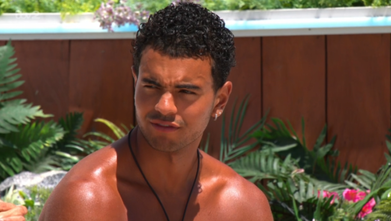 A man with curly dark hair and a serious expression, wearing a black necklace and a silver earring.