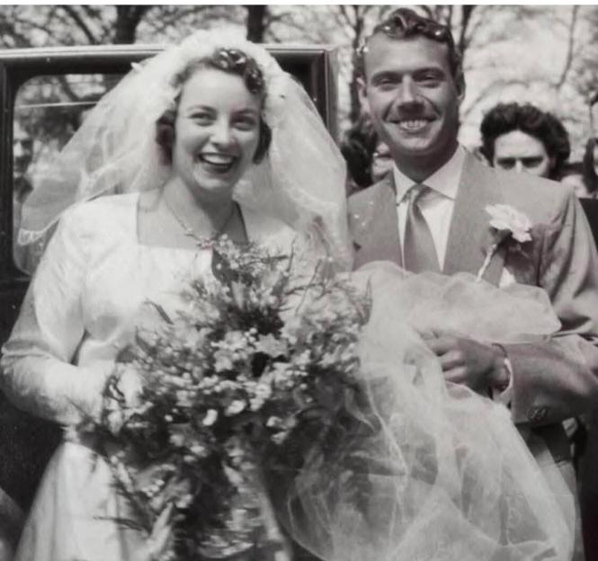 A black and white photo of Joyce, a bride in a white dress and veil, holding a bouquet, and her groom, in a suit, both smiling.