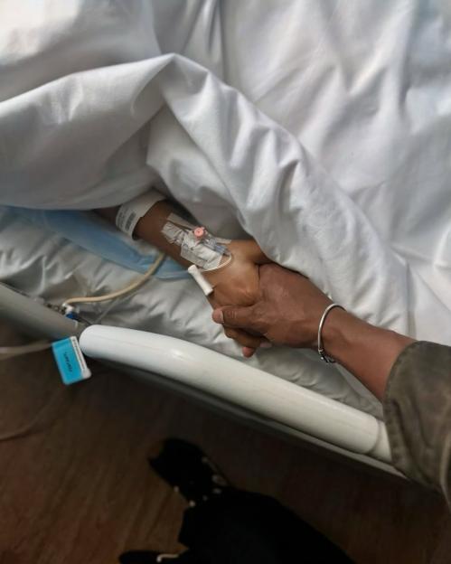 A hand with an IV drip holds another hand, under a white hospital blanket.