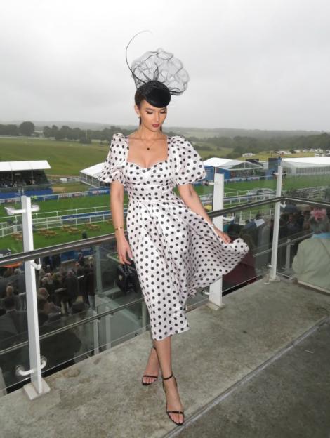 A woman in a white polka-dot dress and black fascinator stands on a balcony overlooking a horse racing track.