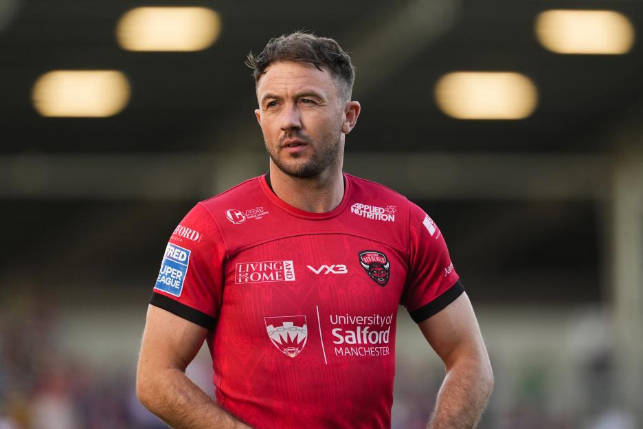 Ryan Brierley, a rugby player in a red "Salford Red Devils" jersey, looks into the distance on the field.