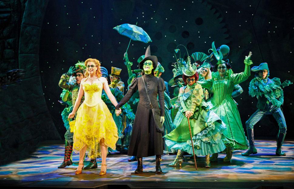 Helen Dallimore (Glinda) in a yellow dress and Idina Menzel (Elphaba) with green skin and a black dress holding hands with other performers in green costumes on stage.