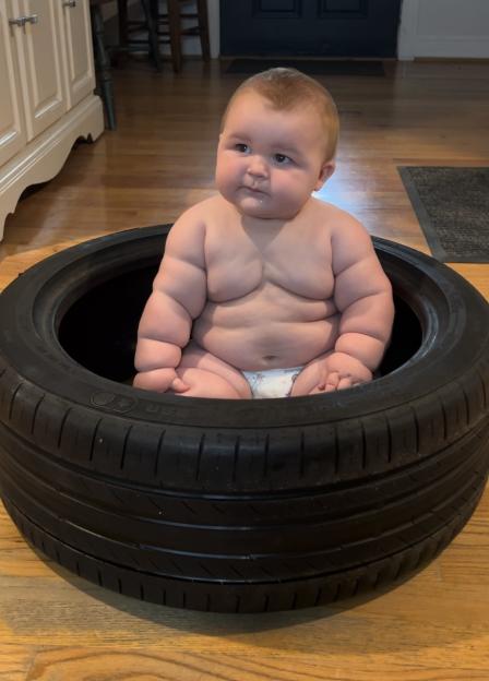 A very large baby in a diaper sitting inside a car tire on a wooden floor.