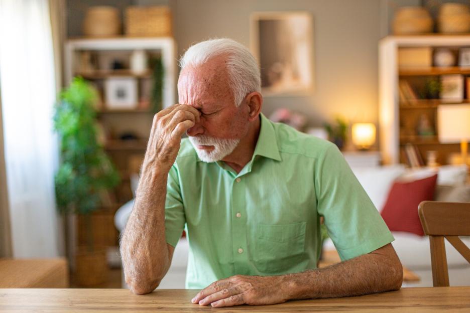 A senior man with a white beard and hair, wearing a green short-sleeved shirt, sitting at a wooden table, suffering from a strong headache and touching his forehead.