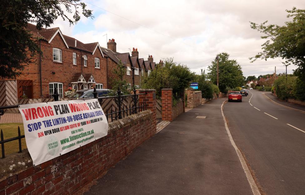 A banner protesting the asylum center in Linton-on-Ouse, reading "WRONG PLAN WRONG PLACE. STOP THE LINTON-ON-OUSE ASYLUM CENTRE. AN OPEN DOOR ASYLUM CENTRE OF 1500 IN A VILLAGE OF 700".
