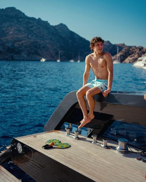 Young man in swim shorts sitting on the back of a boat.