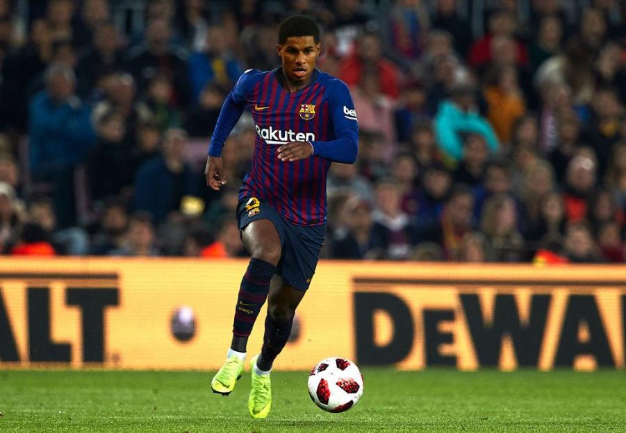 Football player Marcus Rashford in a blue and red striped jersey with Rakuten sponsor, dribbling a soccer ball on a green field.