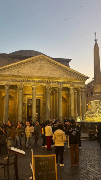 The Pantheon at dusk, with people gathered in front and a sign for happy hour in the foreground.