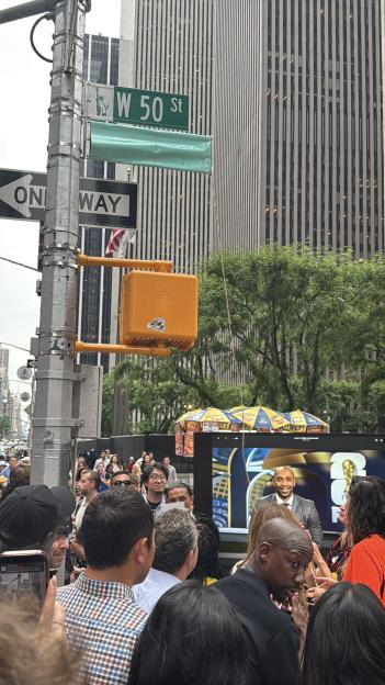 A crowd of people on W 50th St, New York City, gather to watch Thierry Henry on a large outdoor screen.