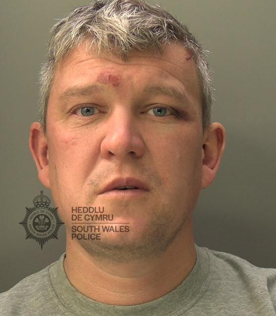A mugshot of Daniel Morgan with minor injuries to his forehead and a black eye, with the South Wales Police emblem partially visible.