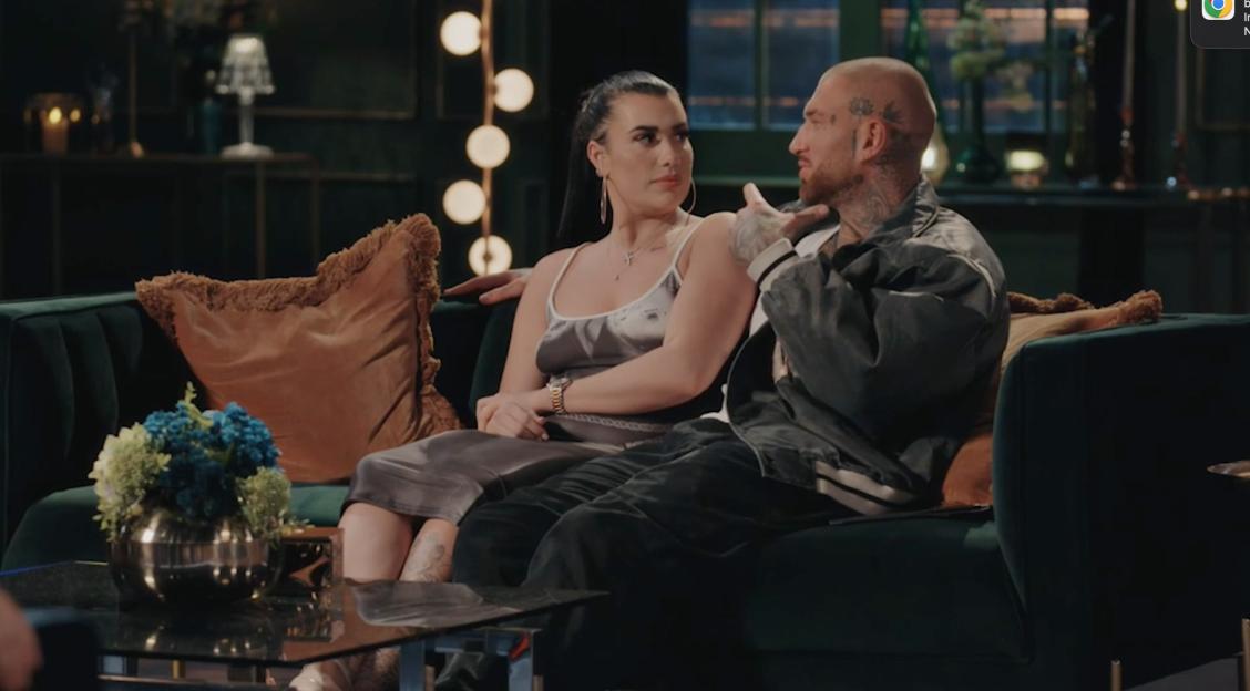 Joe and Maeve sitting together on a green couch, with Joe talking and gesturing with his hand.