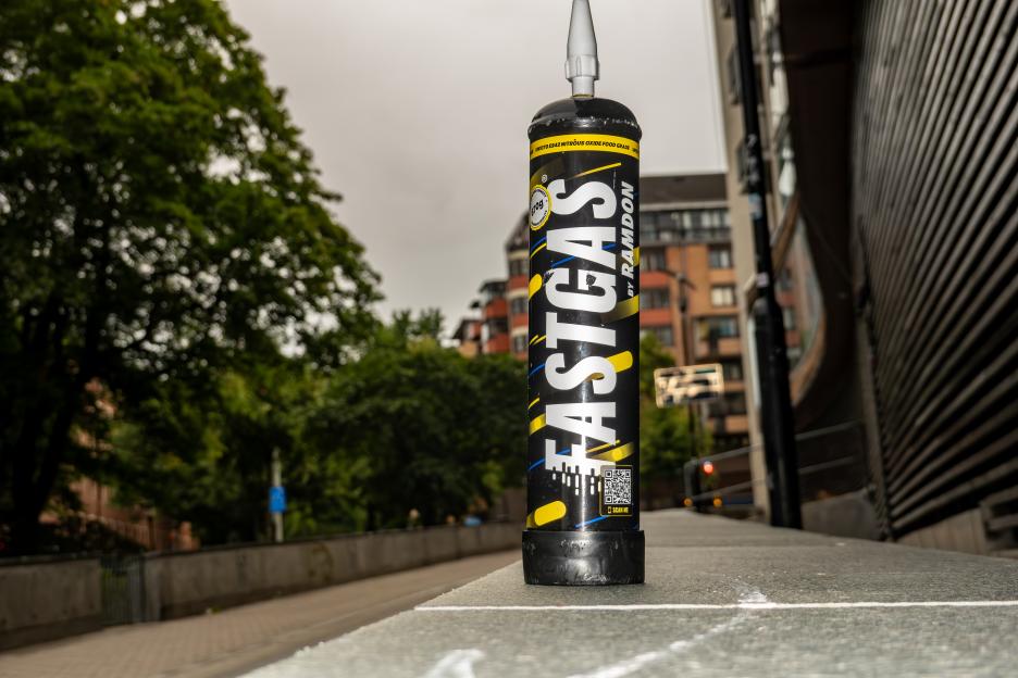A black metal "Fastgas by Ramdon" nitrous oxide canister sitting on a concrete ledge in a city.