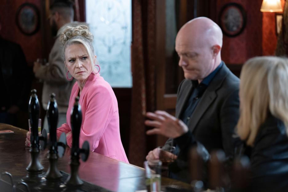 Linda Carter in a pink blazer looks annoyed at Max Branning, who is talking to Cindy Beale at a bar.