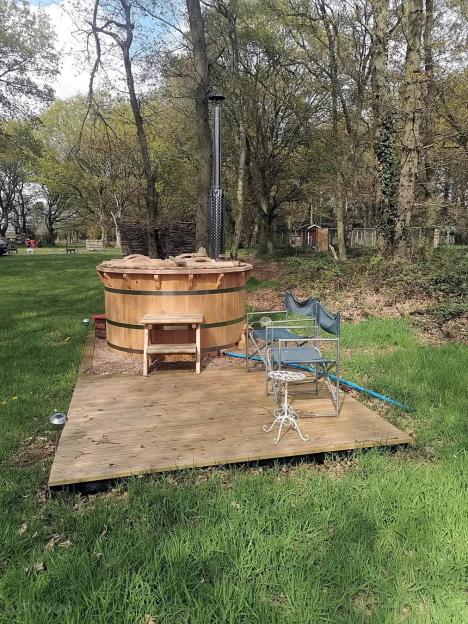 A wooden hot tub with a tall metal chimney on a wooden deck in a grassy area with trees.