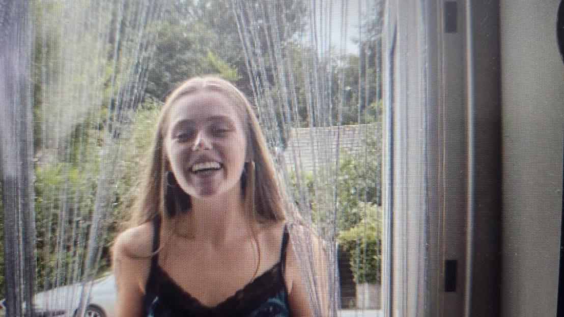 A young woman with long hair smiling and looking at the camera through a streaked window, with greenery and a car visible outside.