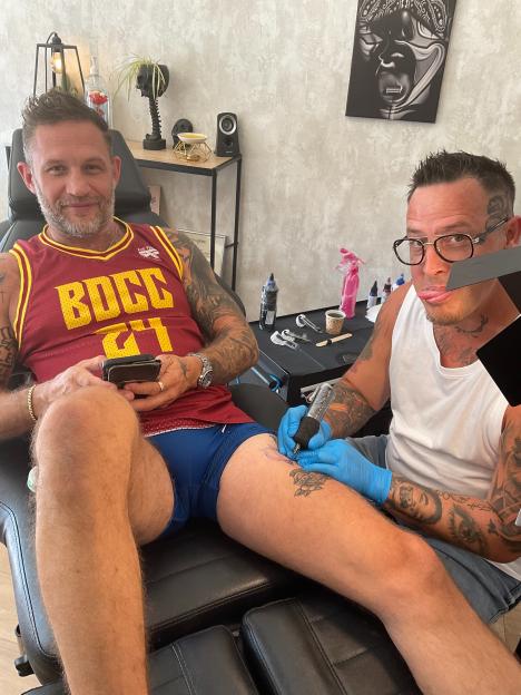 A man in a red jersey getting a tattoo on his upper thigh from a tattoo artist.