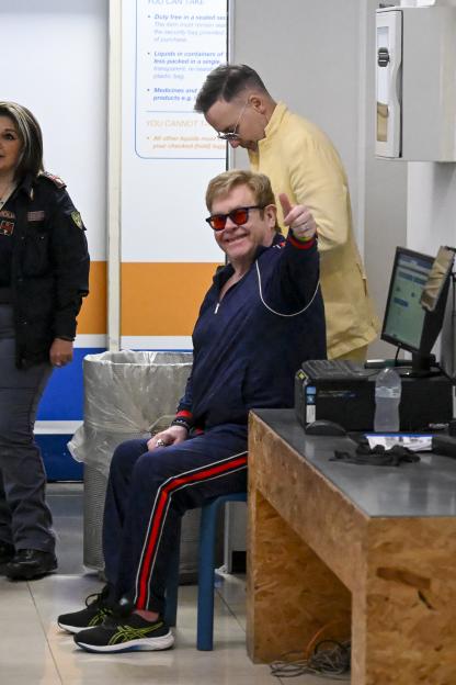Elton John, wearing a navy tracksuit and red sunglasses, gives a thumbs up while sitting, with David Furnish standing behind him.