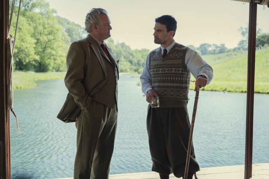 Two men in formal attire stand by a lake, one holding a glass and a walking stick.
