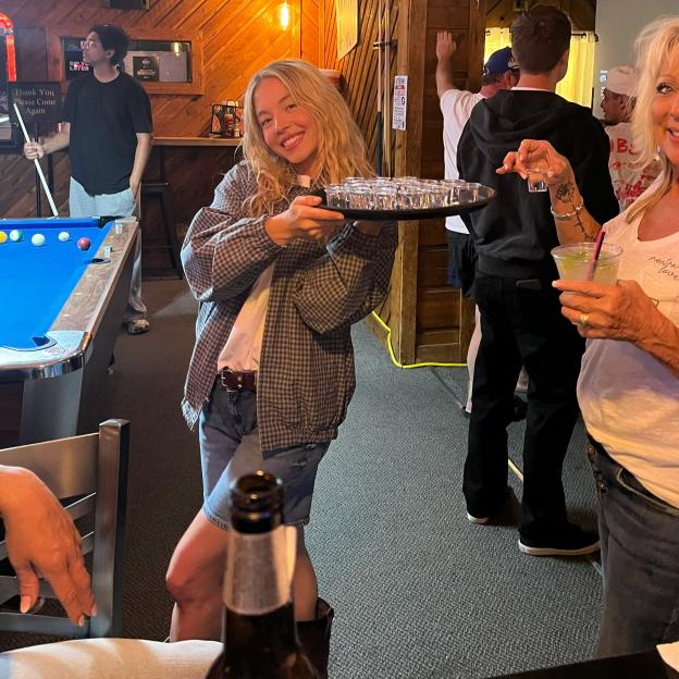 Sydney Sweeney holding a tray of shot glasses in a bar.