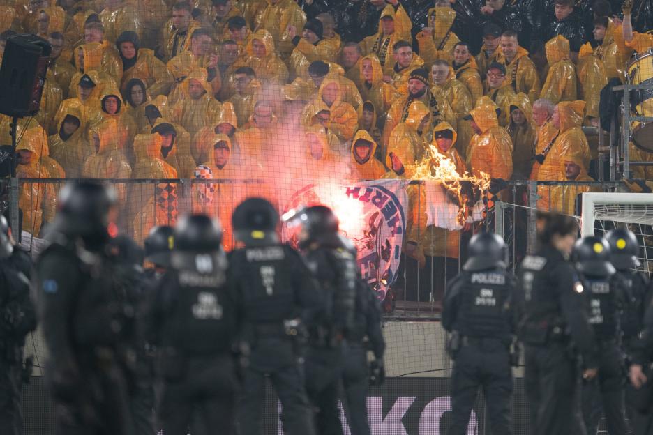Soccer fans in yellow ponchos with pyrotechnics and burning banners, with police in riot gear in the foreground.
