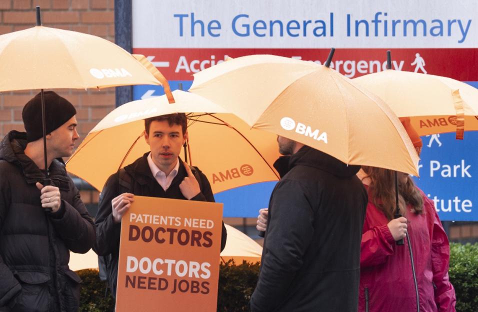 People holding orange umbrellas and a sign that reads "PATIENTS NEED DOCTORS, DOCTORS NEED JOBS" in front of a hospital sign.