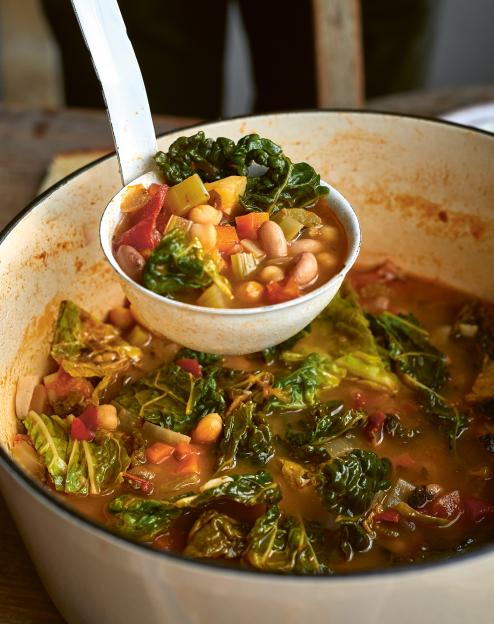 A white ladle serving minestrone soup from a large white pot.