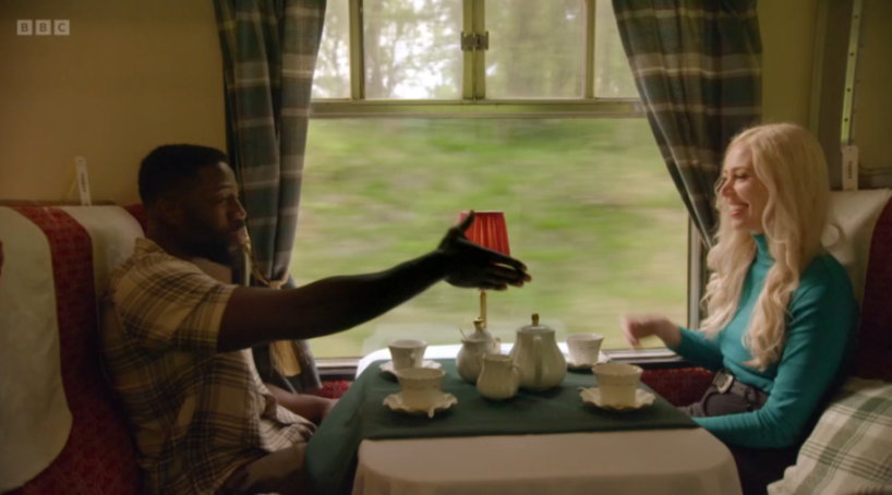 Ellie and Ross on a train, sitting at a table with tea service.