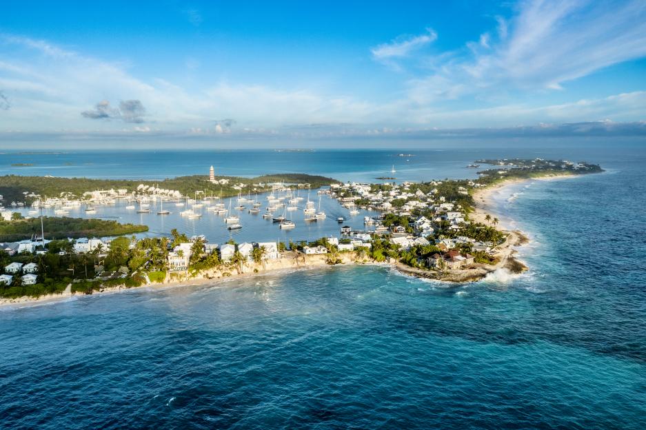 Aerial view of Hope Town, Great Abaco Island, Bahamas with many yachts anchored in the harbor and a striped lighthouse on the land.