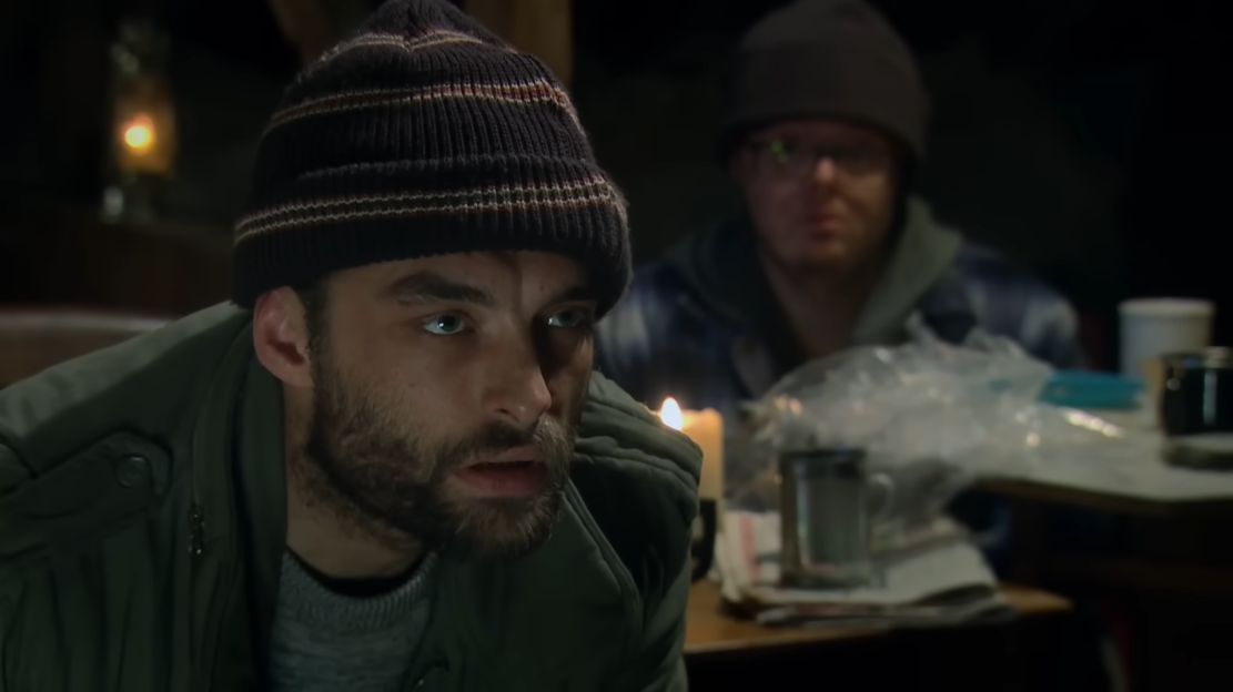 A man with a beard and knit cap looks forward with a concerned expression, another man blurred in the background, a candle and mess on a table.