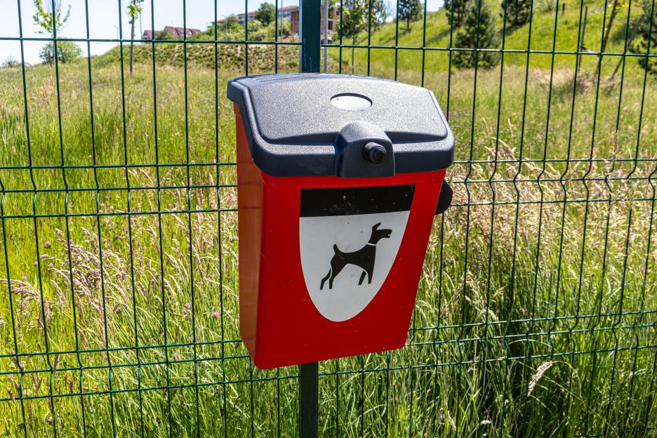 Red and black dog waste bag dispenser with a dog icon on it, attached to a green fence with tall grass and houses in the background.