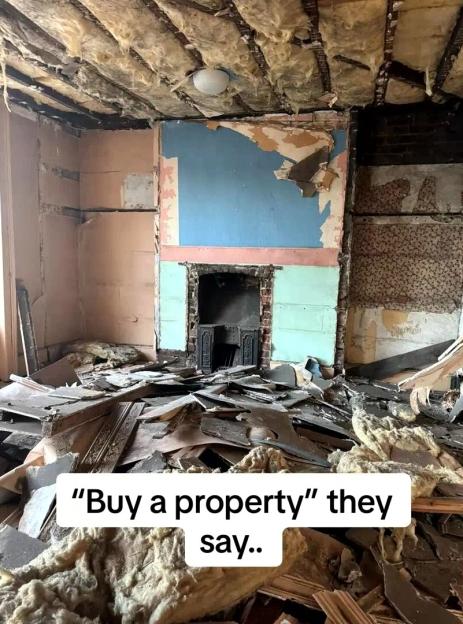 A derelict room with a crumbling ceiling, exposed insulation, peeling wallpaper, a fireplace, and debris covering the floor.