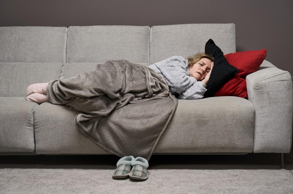 Caucasian woman sleeping on a sofa, covered with a blanket, with two pillows under her head, and slippers on the floor.